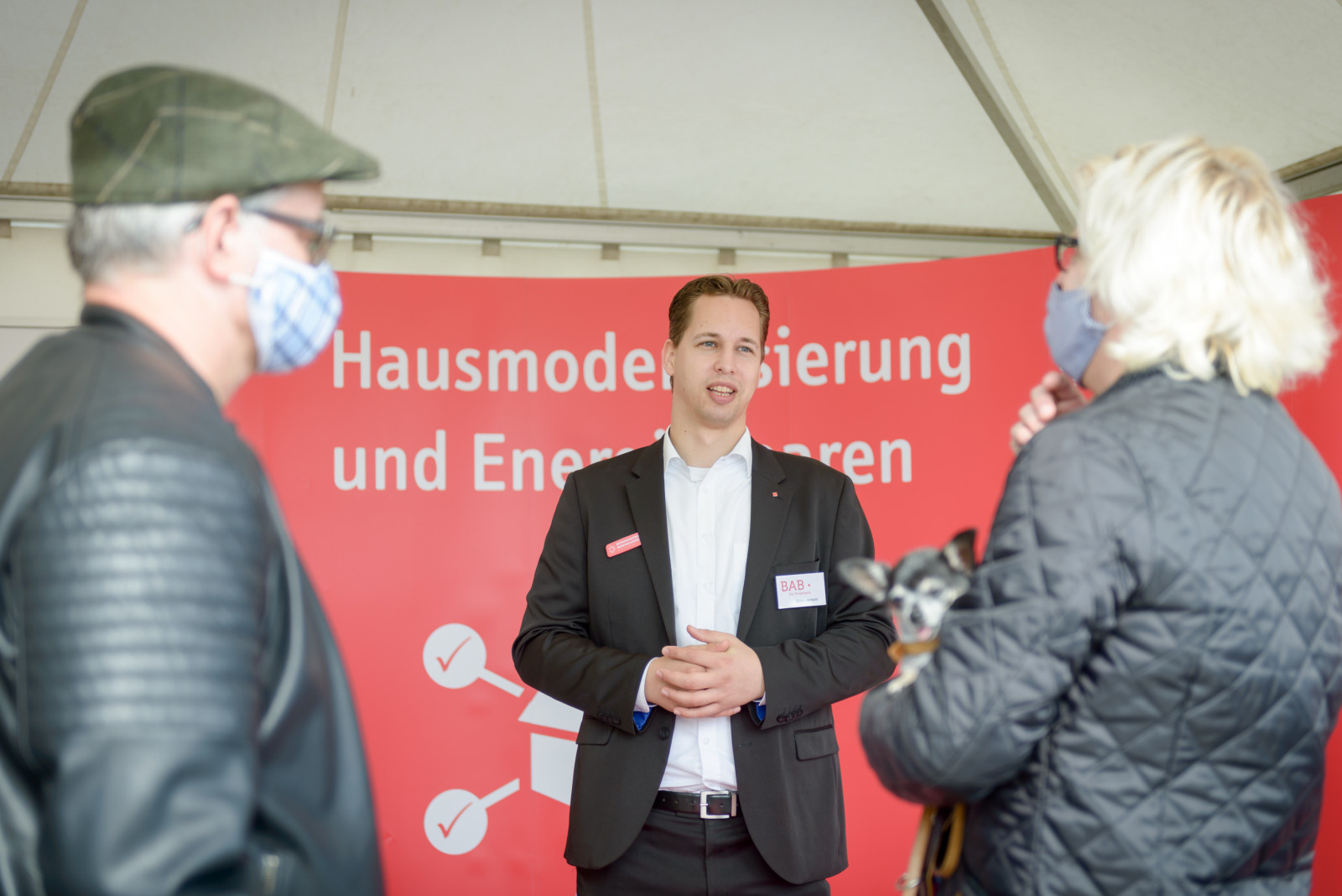 Älteres Ehepaar steht mit Mund-Nasen-Schutz vor Finanzberater Björn Jantzen von der Bremer Aufbau-Bank. Im Hintergrund ist eine Messewand von Bremer Modernisieren.