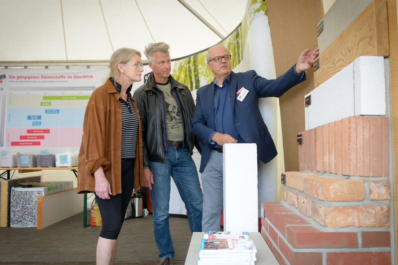 Energieexperte Heinfried Becker steht mit einem Mann und einer Frau neben einem Ausstellungsstück, das mehrere Außenwände zeigt und berät zu unterschiedlichen Dämmmöglichkeiten.